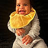 baby, child, smiling, yellow_bib, grey_clothing, portrait, happy, cute, infant, sitting, hands, face, curly_hair, indoor, closeup, young_child, expression, bright, warm, adorable