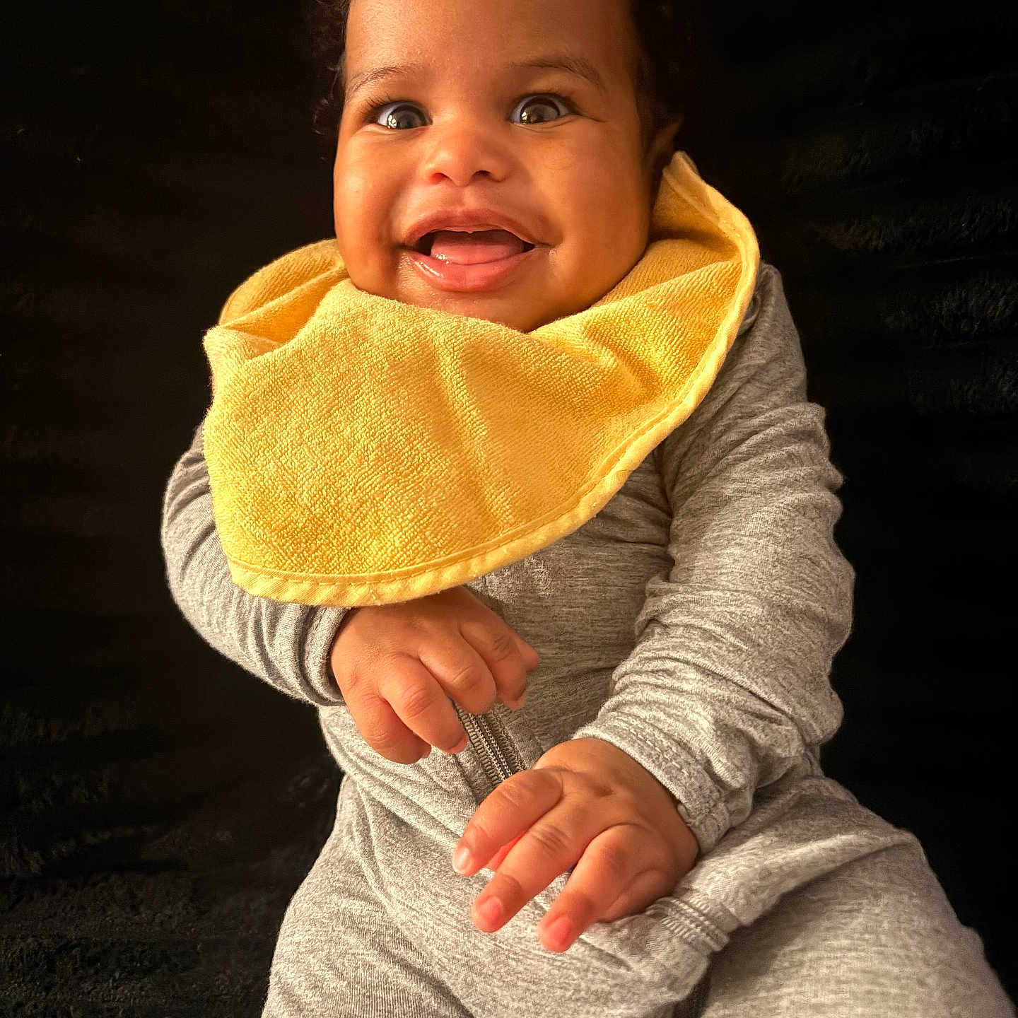 Shanan joined the competition — help win amazing prizes! baby, child, smiling, yellow_bib, grey_clothing, portrait, happy, cute, infant, sitting, hands, face, curly_hair, indoor, closeup, young_child, expression, bright, warm, adorable