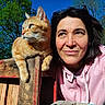 animal, blue_sky, casual_clothing, cat, closeup, daylight, fence, greenery, hoodie, looking_away, nature, orange_cat, outdoor, pet, portrait, relaxed, sunlight, tree, woman, wood