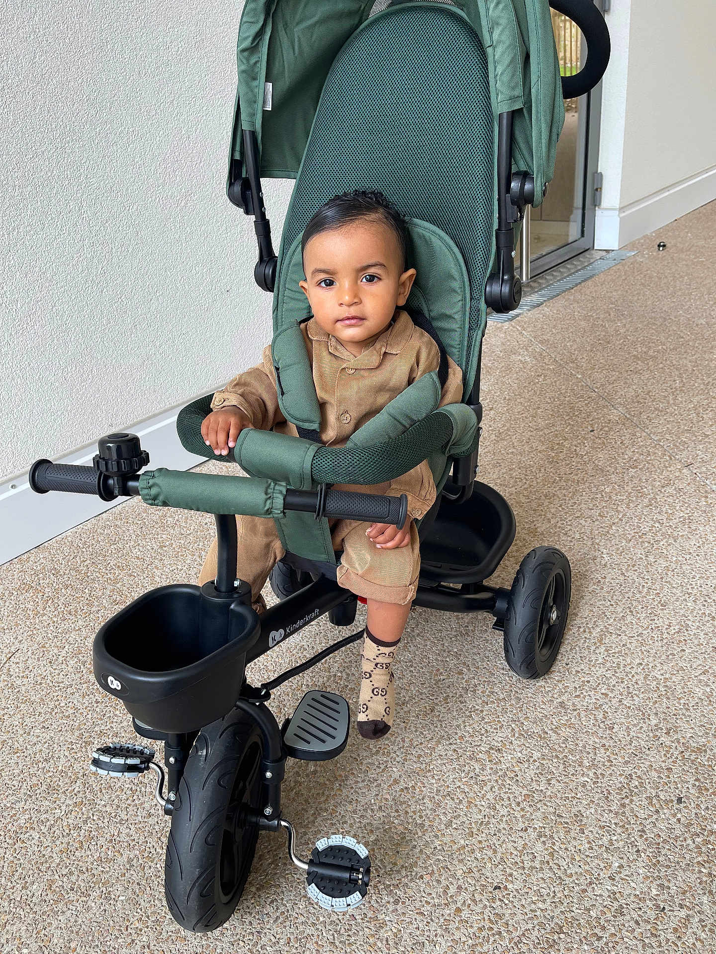 Jaouen a rejoint le concours — aidez-le/la à gagner de superbes lots ! baby, brown_clothing, child, expression, face, floor, green, handlebar, indoor, pedal, person, seatbelt, sitting, socks, stroller, toddler, toy, tricycle, wheel, young_child