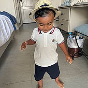 Jaouen a rejoint le concours — aidez-le/la à gagner de superbes lots ! barefoot, bed, casual_clothing, child, desk, floor, hat, indoor, looking_down, navy_shorts, person, room, smiling, standing, tile_floor, toddler, trash_bin, water_bottles, white_polo_shirt, young_child