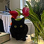 cat, black_cat, tulips, flowers, vase, table, indoor, window, plant_stems, yellow_eyes, domestic_cat, curious, pet, feline, green_leaves, natural_light, houseplant, still_life, cozy, shadow