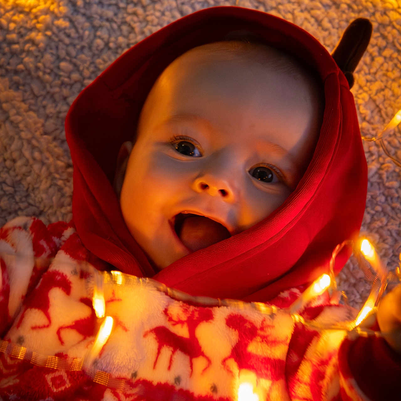 Aëdan a rejoint le concours — aidez-le/la à gagner de superbes lots ! baby, carpet, child, closeup, cozy, cute, face, fairy_lights, festive_sweater, happy, holiday_theme, indoors, infant, playful, portrait, red_hood, reindeer_pattern, smiling, warm_lighting, winter_clothing