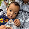 baby, child, infant, portrait, face, wide_eyes, serious_expression, outdoor, porch, adult, parent, hoodie, sweater, snack, food, plastic_bag, hands, family, daylight, casual_photo