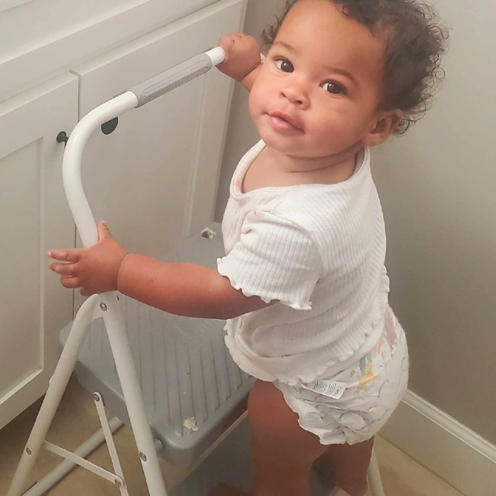 Orunmila joined the competition — help win amazing prizes! barefoot, cabinet, child, curious, curly_hair, diaper, expression, floor, holding, home, indoor, kitchen, light_skin, looking_back, portrait, standing, step_ladder, toddler, white_shirt, young_child