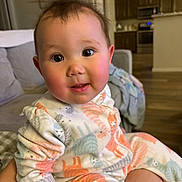 Jovie is registered to the contest to win money with this photo: baby, child, pajamas, animal_print, indoor, living_room, couch, smile, curious, face, person, head, skin, eyes, cheeks, cozy, furniture, floor, home, young_child