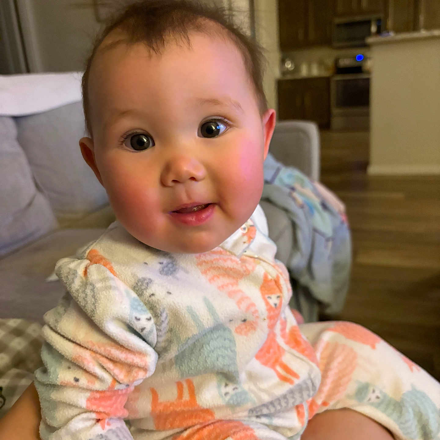 Jovie is registered to the contest to win money with this photo: baby, child, pajamas, animal_print, indoor, living_room, couch, smile, curious, face, person, head, skin, eyes, cheeks, cozy, furniture, floor, home, young_child