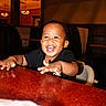 toddler, child, smiling, happy, table, restaurant, indoor, booth, furniture, person, black_shirt, high_chair, lighting, face, hands, joy, cute, baby, seat, hair
