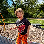 Uhtred is registered to the contest to win money with this photo: child, boy, smiling, playground, slide, orange_pants, pumpkin_pattern, black_tshirt, lightning_mcqueen, outdoor, sunny, trees, grass, shoes, happy, young, face, clothing, play, daytime