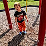 Uhtred is registered to the contest to win money with this photo: child, playground, wood_chips, orange_pants, pumpkin_pattern, black_tshirt, lightning_mcqueen, red_poles, shade, grass, outdoor, sunlight, shoes, person, curious, standing, play_equipment, trees, daytime, casual_clothing