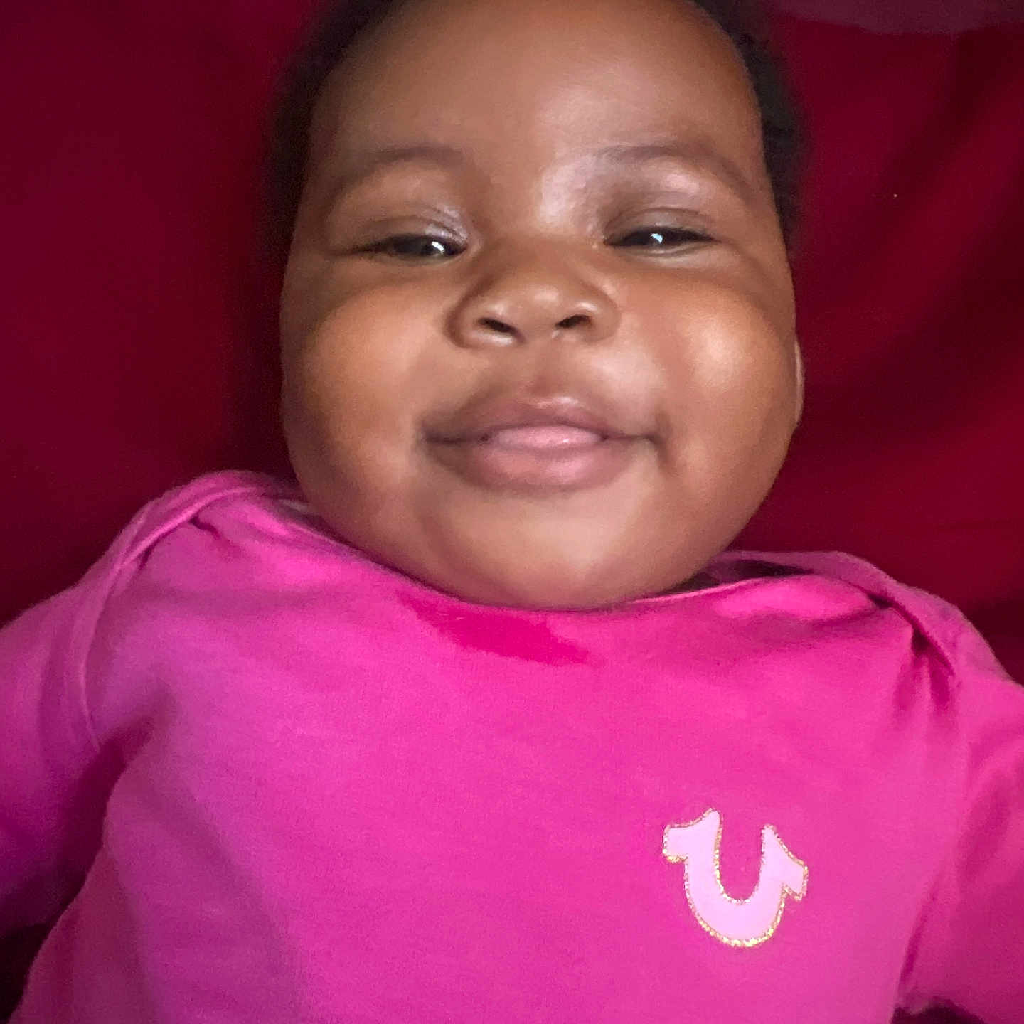 Kamahni is registered to the contest to win money with this photo: baby, child, closeup, clothing, cute, eyebrows, eyes, face, happy, head, indoors, infant, mouth, person, pink_shirt, portrait, red_background, skin, smiling, young_child