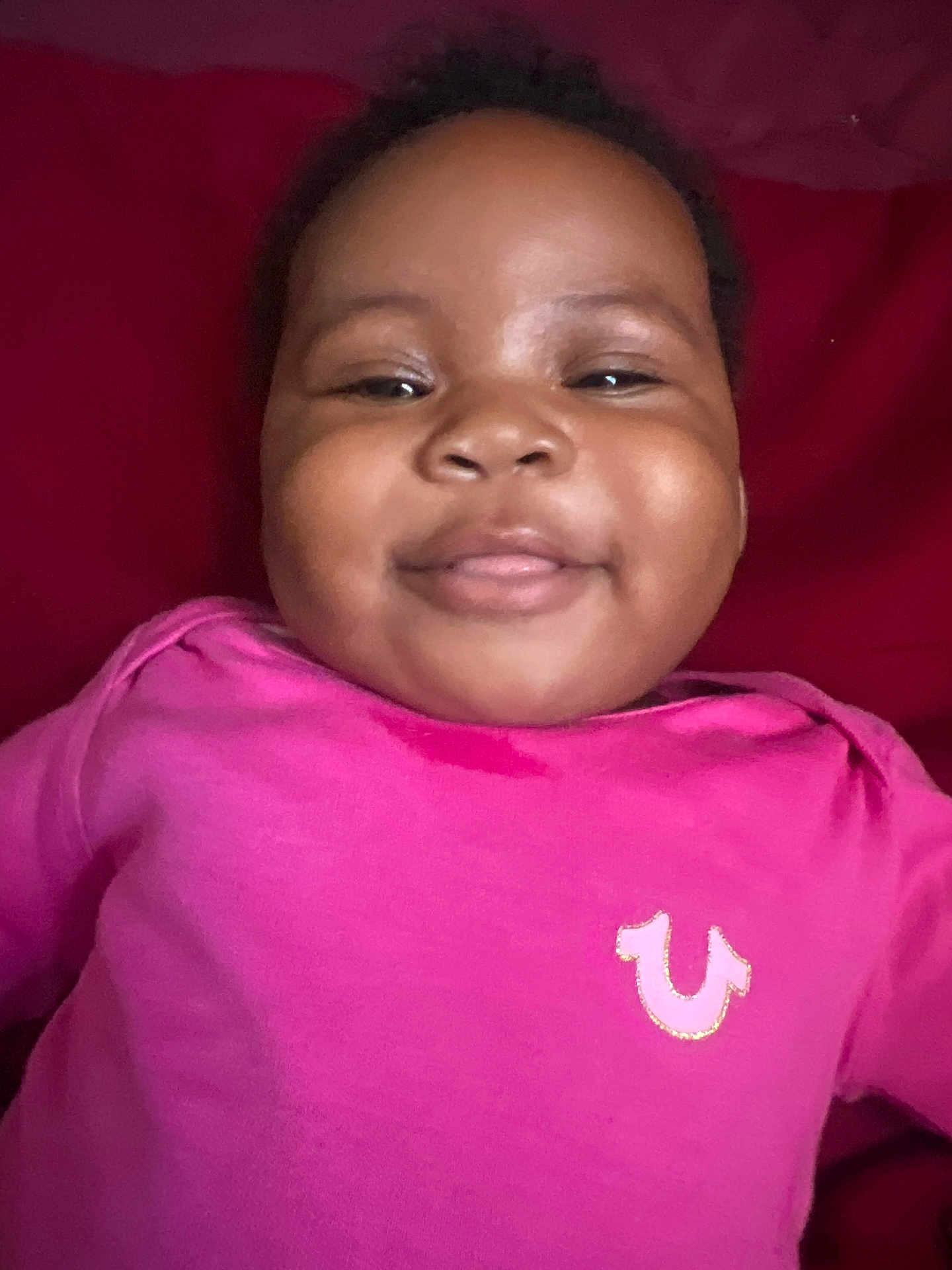 Kamahni is registered to the contest to win money with this photo: baby, smiling, child, pink_shirt, face, happy, infant, portrait, cute, red_background, person, young_child, clothing, closeup, skin, head, eyebrows, eyes, mouth, indoors