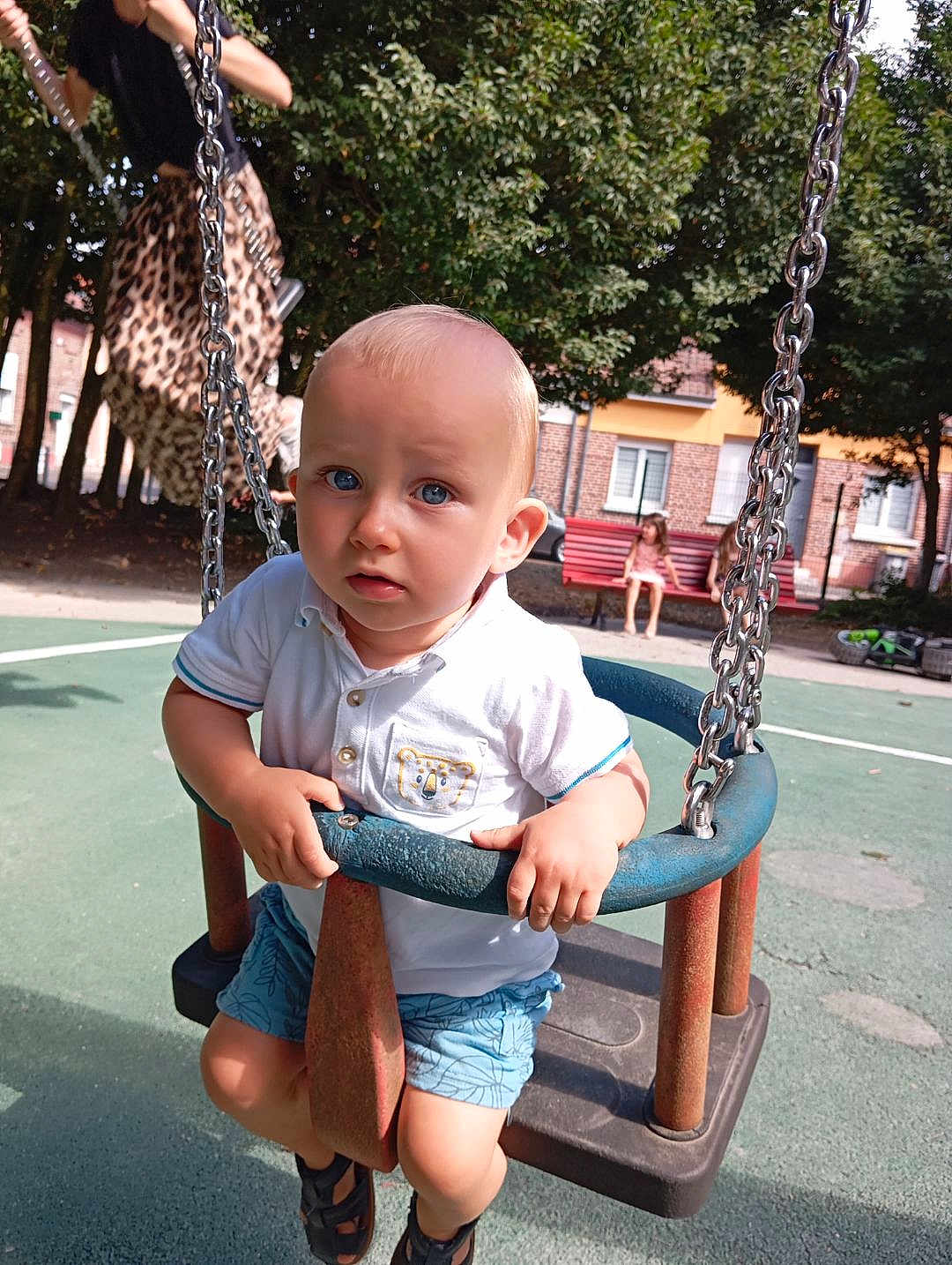 Eliott a rejoint le concours — aidez-le/la à gagner de superbes lots ! toddler, child, swing, playground, outdoor, summer, tree, bench, park, blue_eyes, shorts, white_shirt, sandals, chain, metal, daylight, curious, expression, seat, person