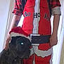 man, dog, santa_hat, apron, plaid_pajamas, indoor, holiday, christmas, pet, beard, tattoo, black_dog, smile, standing, doorway, costume, festive, face, animal, human