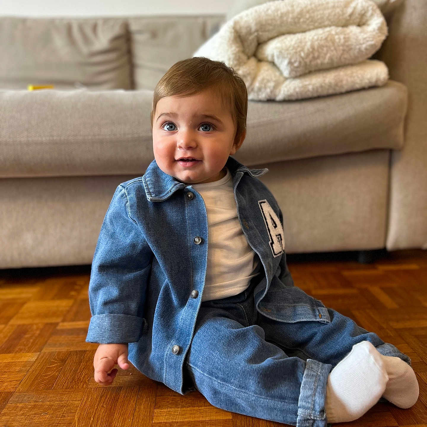 Arié participe au concours pour gagner de l'argent avec cette photo : baby, blanket, casual, child, couch, cozy, cute, denim_jacket, denim_pants, fashion, flooring, home, indoor, portrait, sitting, smile, toddler, white_shirt, white_socks, wooden_floor