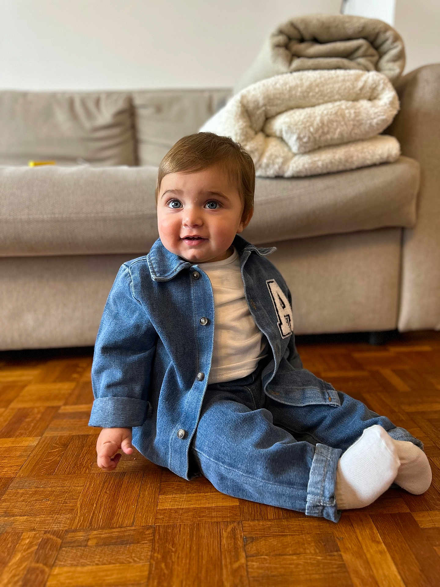 Arié participe au concours pour gagner de l'argent avec cette photo : baby, blanket, casual, child, couch, cozy, cute, denim_jacket, denim_pants, fashion, flooring, home, indoor, portrait, sitting, smile, toddler, white_shirt, white_socks, wooden_floor