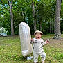 Parvin is registered to the contest to win money with this photo: toddler, child, balloon, number_one, overalls, cap, grass, tree, forest, park, nature, pine_cone, outdoor, happy, smiling, walking, daylight, footwear, clothing, person