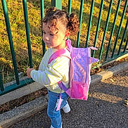 Hayden participe au concours pour gagner de l'argent avec cette photo : child, curly_hair, pigtails, backpack, pink, jeans, white_sneakers, striped_sweater, outdoor, side_view, fence, green_fence, sunlight, shadow, pavement, grass, trees, park, daytime, casual_clothing