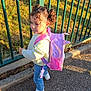 child, curly_hair, pigtails, backpack, pink, jeans, white_sneakers, striped_sweater, outdoor, side_view, fence, green_fence, sunlight, shadow, pavement, grass, trees, park, daytime, casual_clothing