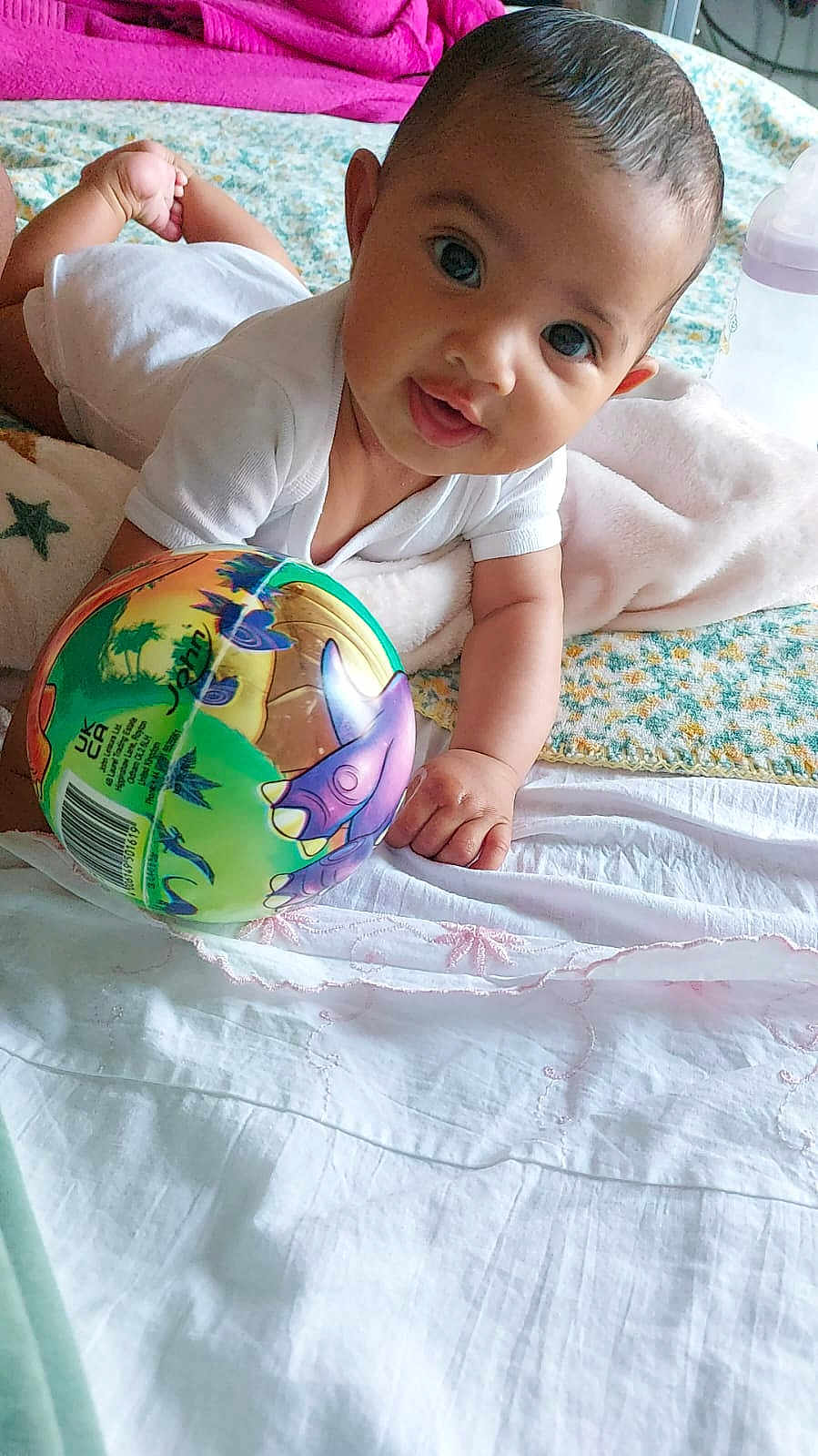 Rose participe au concours pour gagner de l'argent avec cette photo : baby, infant, child, onesie, toy, ball, dinosaur, bed, blanket, playful, cute, indoors, person, smiling, hand, face, head, lying_down, colorful, soft