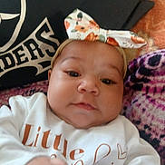 Emiliana joined the competition — help win amazing prizes! baby, child, infant, face, headband, bow, clothing, white_shirt, hand, pillow, text, blanket, pattern, closeup, portrait, indoors, cute, young, person, skin