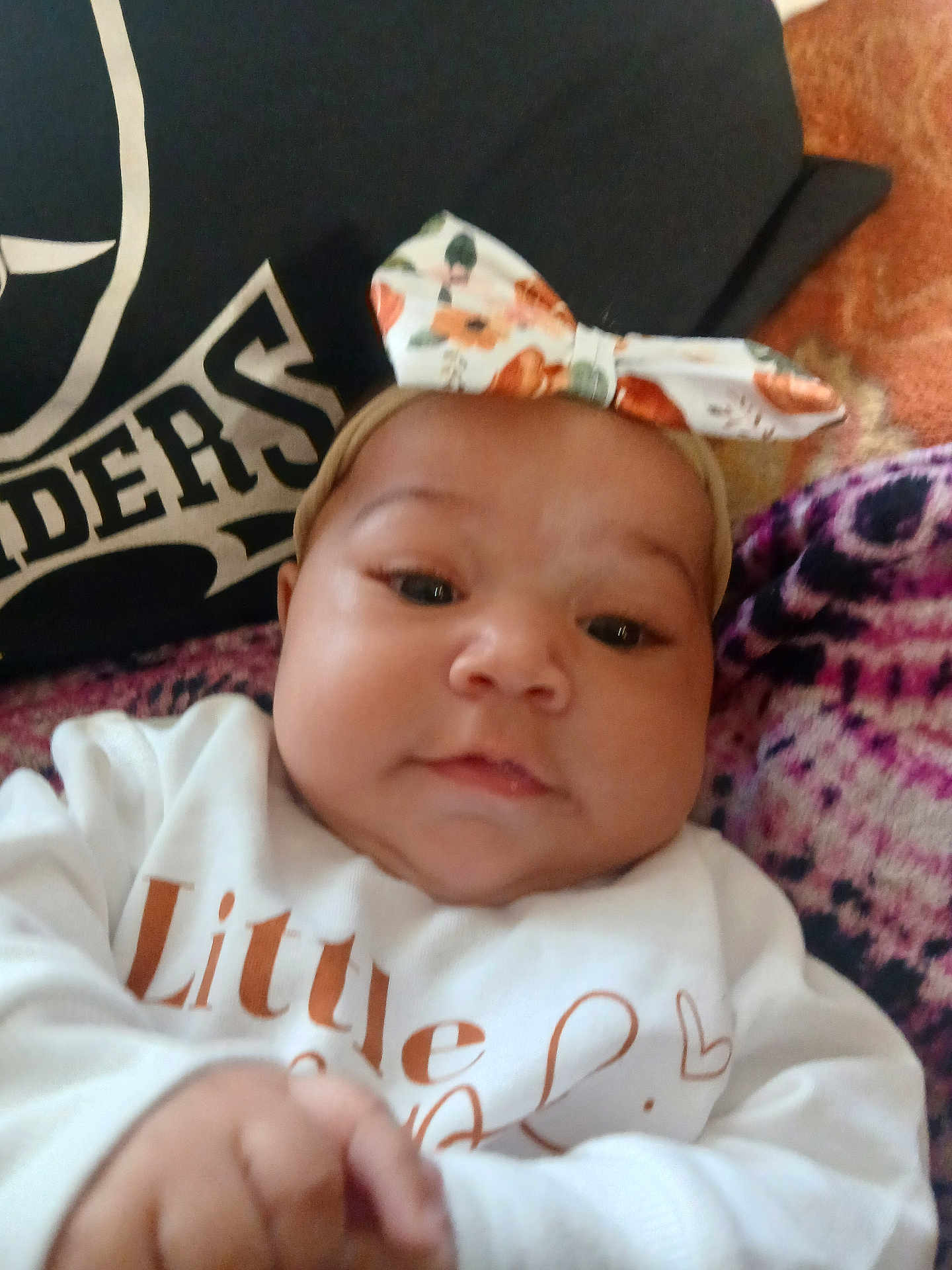 Emiliana joined the competition — help win amazing prizes! baby, child, infant, face, headband, bow, clothing, white_shirt, hand, pillow, text, blanket, pattern, closeup, portrait, indoors, cute, young, person, skin