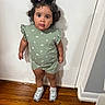 toddler, child, girl, standing, green_outfit, pigtails, curly_hair, white_wall, wooden_floor, sneakers, socks, indoors, casual_clothing, portrait, cute, young_child, person, full_body, expression, looking_up