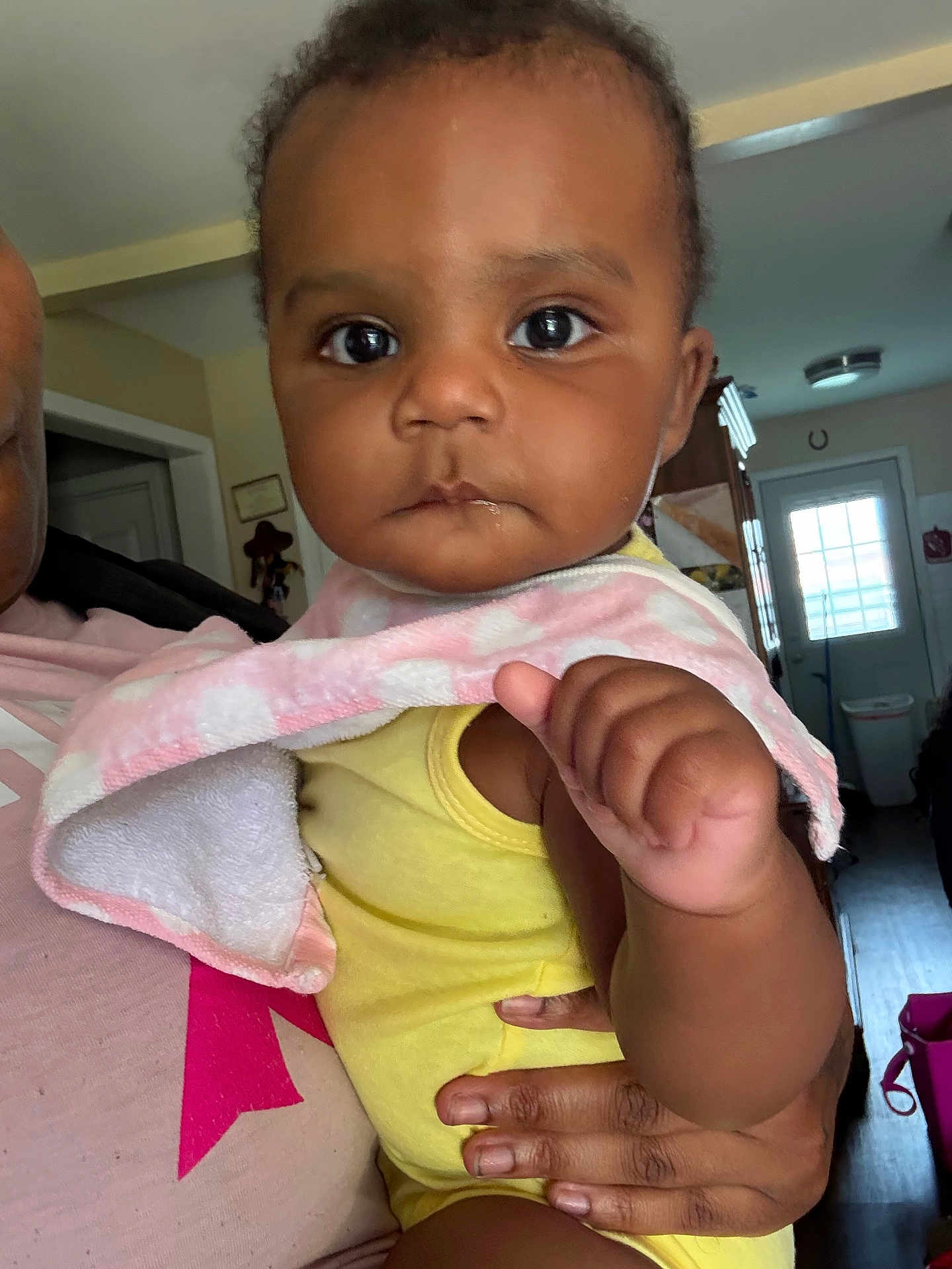 Journee joined the competition — help win amazing prizes! baby, child, infant, face, hand, yellow_clothing, pink_bib, indoor, person, holding, home, room, door, natural_light, portrait, expression, skin, furniture, adult_hand, serious