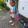 child, girl, backpack, outdoor, concrete, house, door, window, hair_beads, sneakers, shorts, tshirt, minnie_mouse, plants, sidewalk, daylight, expression, standing, curious, colorful