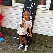 Kyrah joined the competition — help win amazing prizes! child, birthday, girl, peace_sign, purple_beads, sneakers, window, reflection, outdoor, concrete, sign, white_shirt, black_shorts, smiling, person, celebration, balloons, plants, hair_beads, casual_clothing