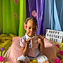 child, easter_eggs, colorful, braided_hair, beads, pink_blanket, playful_pose, smiling, indoor, decorations, lightbox_sign, pajama_shirt, white_shorts, curtains, paw_prints, yellow_eggs, purple_eggs, pink_eggs, gold_egg, portrait