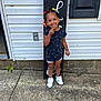 child, toddler, smiling, standing, colorful_hair_beads, black_shirt, white_shorts, white_sneakers, concrete, house, window, shutter, outdoor, happy, person, playful, curly_hair, front_door, sidewalk, daylight