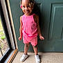 child, girl, smiling, standing, indoors, door, pink_clothing, shorts, tank_top, white_sneakers, braided_hair, beads, floor, window, natural_light, happy, person, portrait, casual_wear, cute