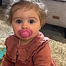 Romy a rejoint le concours — aidez-le/la à gagner de superbes lots ! baby, big_eyes, brown_hair, carpet, child, curious_look, cute, dress, floor, furniture, hair_clip, indoor, pacifier, pink_pacifier, portrait, ruffled_collar, rug, sitting, sofa, toy