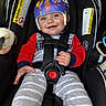 baby, child, helmet, car_seat, smile, pajamas, red_sleeves, striped_pants, plush_toy, safety_harness, cute, infant, indoors, seat_belt, protective_gear, happy, person, closeup, blue_eyes, casual