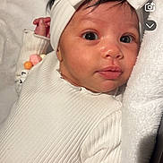 Layana participe au concours pour gagner de l'argent avec cette photo : baby, infant, newborn, headband, bow, white_outfit, ribbed_texture, blanket, towel, beads, bracelet, closeup, portrait, face, eyes, gaze, indoor, soft_light, cozy, crib