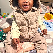 Jacari joined the competition — help win amazing prizes! toddler, child, baby, hooded_vest, smile, indoor, bed, blanket, pants, barefoot, happy, cute, person, portrait, cozy, toy, room, clothing, sitting, face