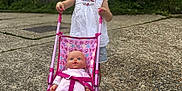 Delilah joined the competition — help win amazing prizes! toddler, child, toy, stroller, baby_doll, outdoor, concrete, fence, grass, greenery, smile, blonde_hair, bows, white_shirt, shorts, play, daylight, person, happy, casual