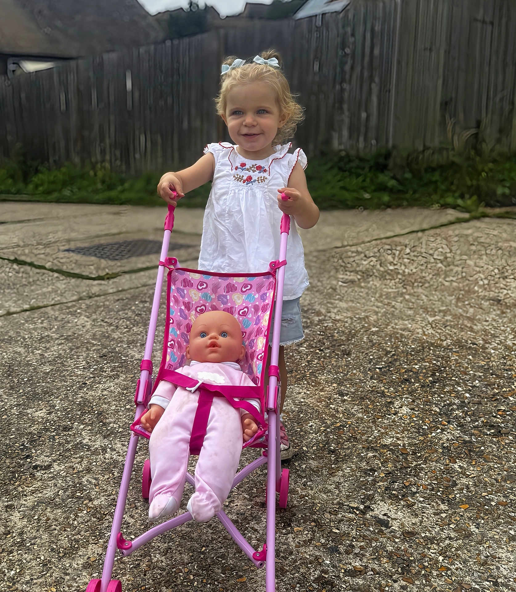 Delilah joined the competition — help win amazing prizes! toddler, child, toy, stroller, baby_doll, outdoor, concrete, fence, grass, greenery, smile, blonde_hair, bows, white_shirt, shorts, play, daylight, person, happy, casual