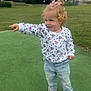 toddler, child, pointing, curly_hair, pink_bows, white_sweater, polka_dots, cartoon_characters, light_blue_jeans, crocs, outdoor, green_field, grass, trees, basketball_court, daylight, smiling, happy, playful, casual_clothing