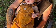 Cruz is registered to the contest to win money with this photo: baby, child, adult, tattoos, overalls, beanie, outdoor, grass, lawn_chair, table, can, sunlight, person, casual_clothing, greenery, relaxation, barefoot, summer, portrait, family