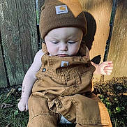 Cruz is registered to the contest to win money with this photo: baby, child, outdoor, grass, wooden_fence, brown_clothing, hat, beanie, overalls, sunlight, shadow, curious, sitting, person, nature, daylight, cute, infant, apparel, casual