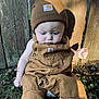 baby, child, outdoor, grass, wooden_fence, brown_clothing, hat, beanie, overalls, sunlight, shadow, curious, sitting, person, nature, daylight, cute, infant, apparel, casual