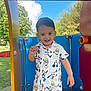 child, toddler, smiling, playground, outdoor, blue_sky, greenery, shirt, shorts, cartoon_print, happy, person, daylight, colorful, play_equipment, cute, young_child, summer_clothing, standing, park