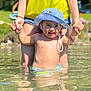 Elyas a rejoint le concours — aidez-le/la à gagner de superbes lots ! toddler, child, water, hat, striped_hat, summer, outdoor, sunlight, smile, happy, adult, holding_hands, yellow_top, denim_shorts, shallow_water, reflection, nature, greenery, play, fun