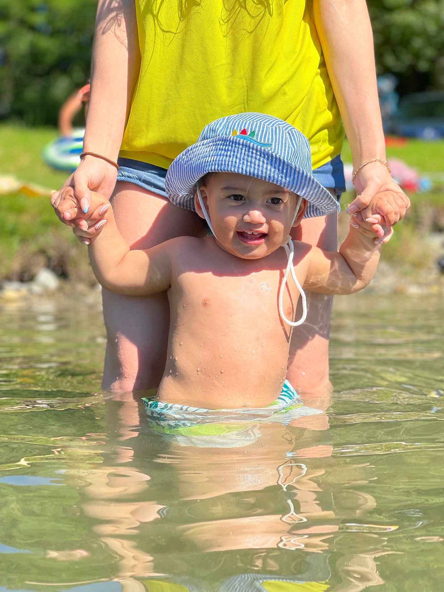 Elyas a rejoint le concours — aidez-le/la à gagner de superbes lots ! toddler, child, water, hat, striped_hat, summer, outdoor, sunlight, smile, happy, adult, holding_hands, yellow_top, denim_shorts, shallow_water, reflection, nature, greenery, play, fun