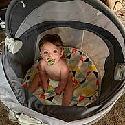 Ridge is registered to the contest to win money with this photo: baby, pacifier, playpen, colorful_mat, geometric_pattern, indoor, carpet, child, sitting, curious, cute, infant, soft_lighting, head, face, brown_hair, diaper, toy_area, furniture, home