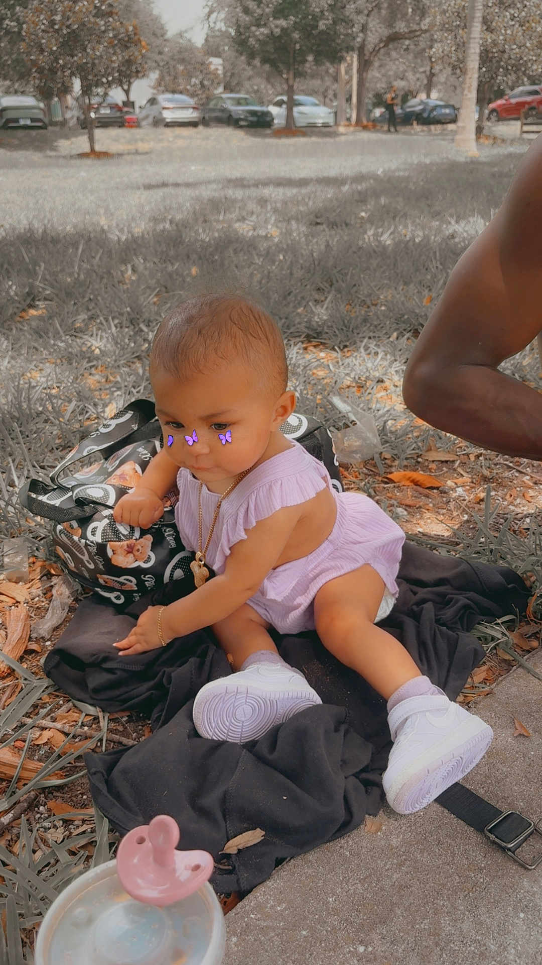 Yazmin is registered to the contest to win money with this photo: baby, child, outdoor, grass, sneakers, purple_outfit, jewelry, pacifier, cup, bag, person, trees, sidewalk, fall_leaves, playing, cute, accessories, sunlight, blurred_background, purple_butterflies