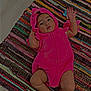 baby, child, pink_costume, dinosaur_outfit, rug, colorful_rug, floor, indoors, cute, infant, looking_up, hand_raised, laying_down, costume, playful, portrait, expression, baby_clothes, soft_texture, young_child