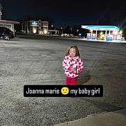 Joanna is registered to the contest to win money with this photo: child, girl, night, parking_lot, gas_station, building, car, jacket, bottle, outdoor, streetlight, pavement, person, clothing, smiling, holding, urban, sidewalk, dark, portrait